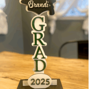 Graduation decoration featuring the name "Brandi," the word "GRAD" in green and white letters, and the year "2025," displayed on a wooden table.