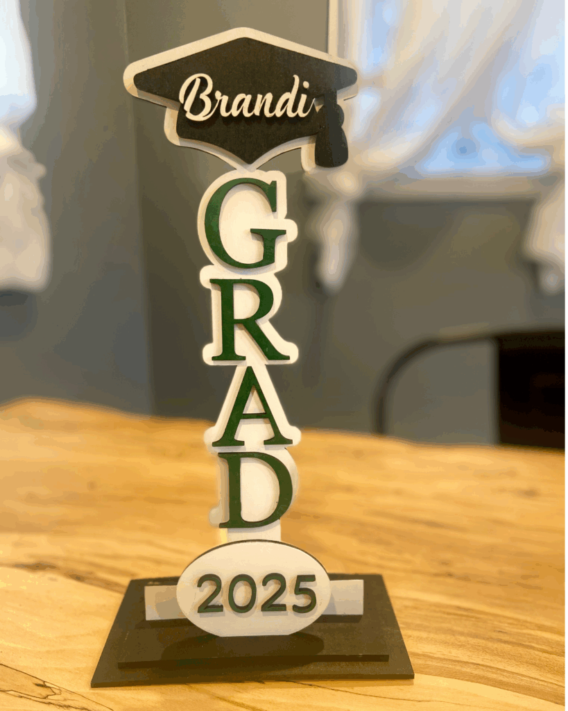 Graduation decoration featuring the name "Brandi," the word "GRAD" in green and white letters, and the year "2025," displayed on a wooden table.