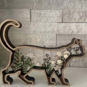 Wooden cat silhouette filled with layered green and floral cutouts, showcasing a decorative design element against a stone wall backdrop.