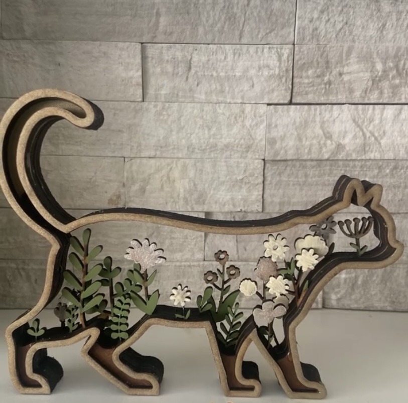 Wooden cat silhouette filled with layered green and floral cutouts, showcasing a decorative design element against a stone wall backdrop.