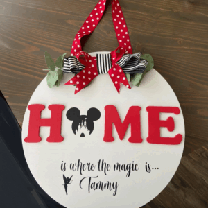 Decorative round sign with red letters spelling "HOME," featuring a Mickey Mouse silhouette, ribbons, and the text "is where the magic is... Tammy."