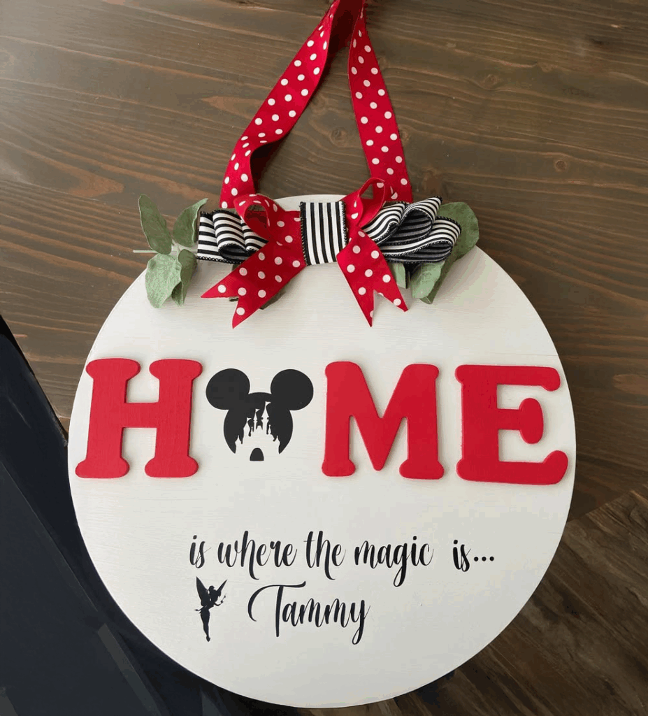 Decorative round sign with red letters spelling "HOME," featuring a Mickey Mouse silhouette, ribbons, and the text "is where the magic is... Tammy."