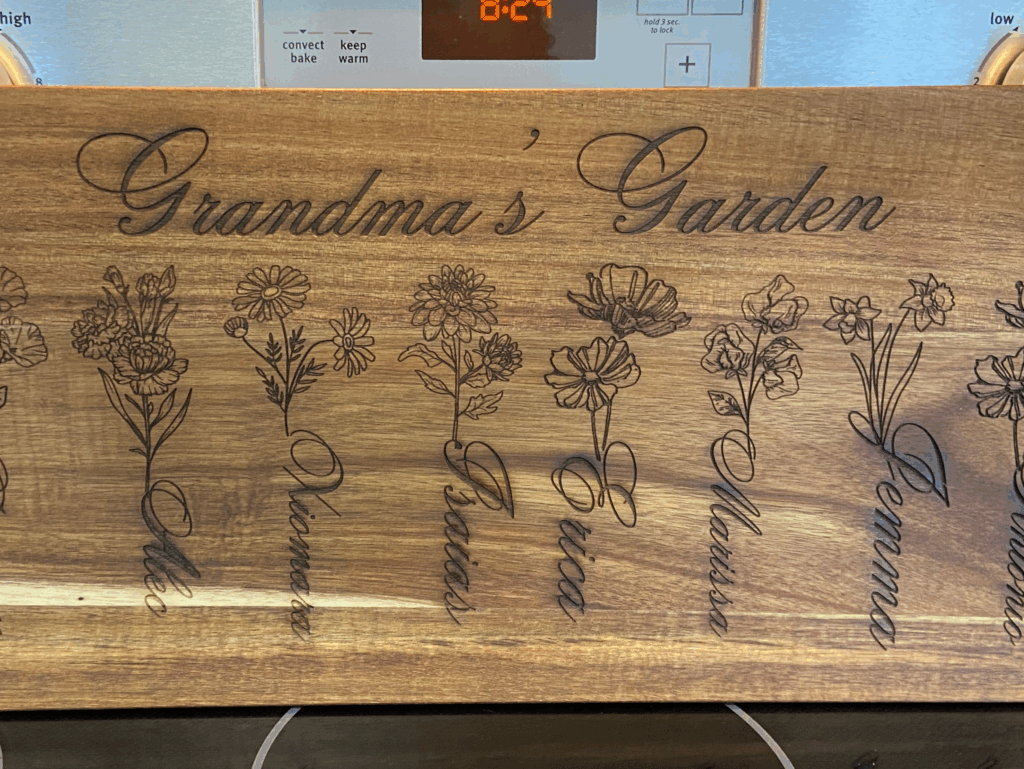 Wooden cutting board engraved with "Grandma's Garden," featuring illustrations of flowers and names: Denver, Elie, Vinas, Erica, Melissa, Gemma, and Antonio.