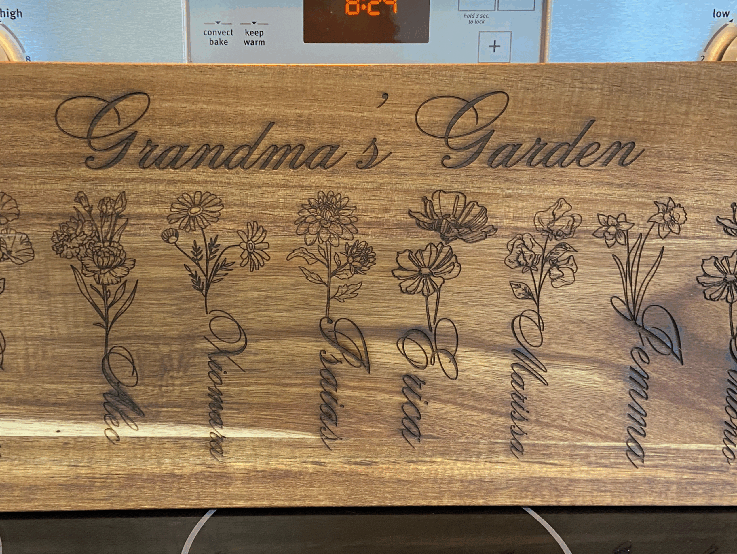 Wooden cutting board engraved with "Grandma's Garden," featuring illustrations of flowers and names: Denver, Elie, Vinas, Erica, Melissa, Gemma, and Antonio.