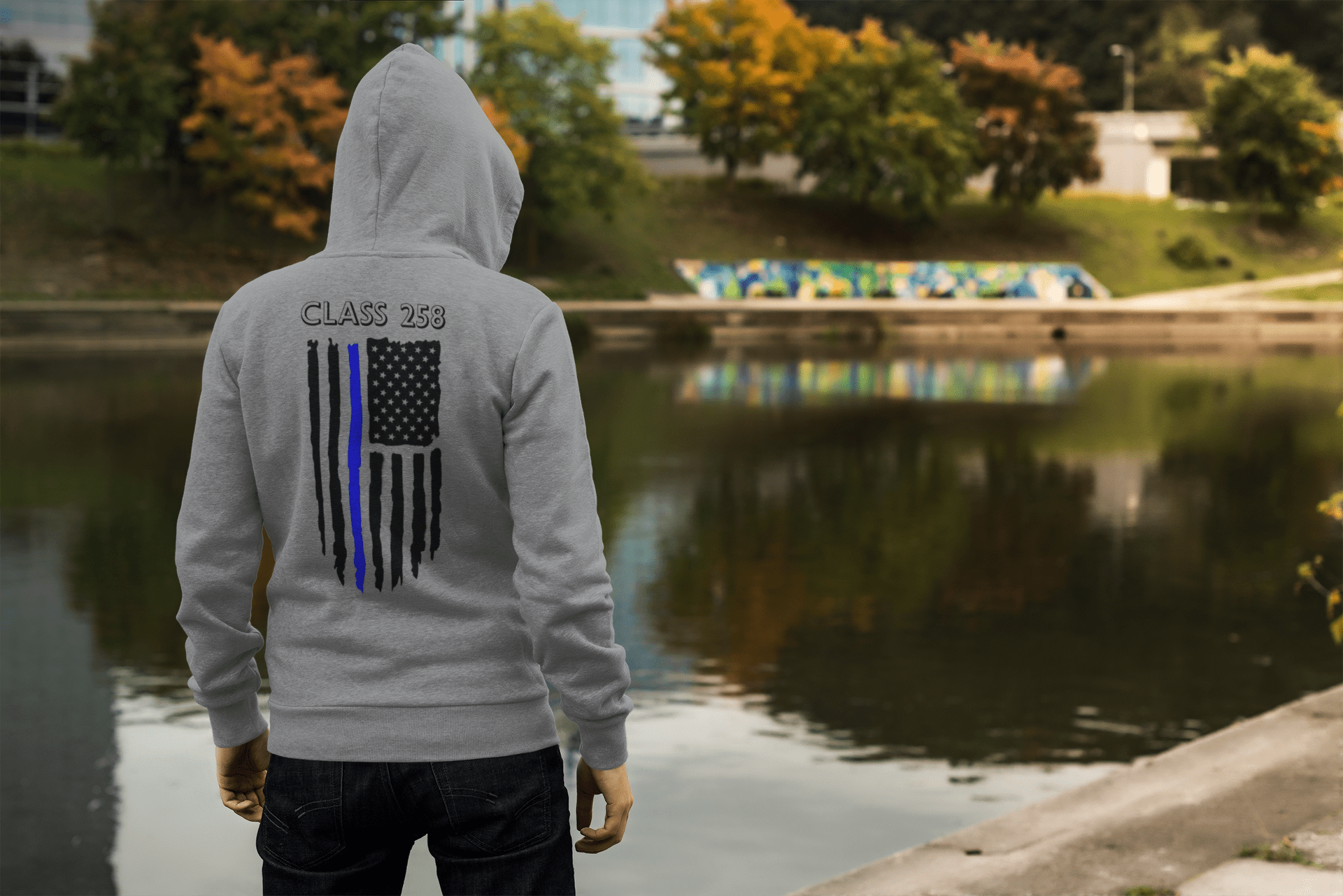A person wearing a gray hoodie with "CLASS 258" and a stylized American flag stands by a tranquil lake, reflecting autumn foliage and graffiti.