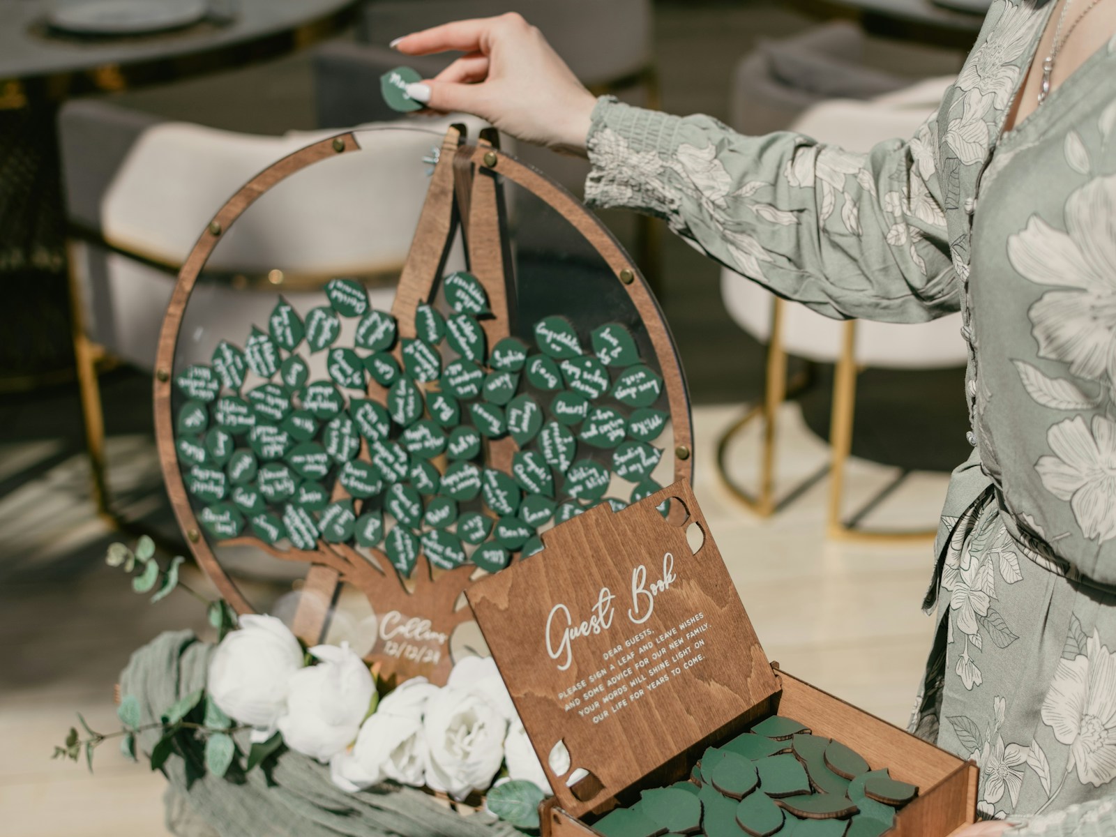 A hand places a green leaf-shaped guestbook token into a wooden display adorned with white roses, inviting guests to write messages.